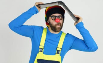 A clown in bright clothes holds a laptop above his head.