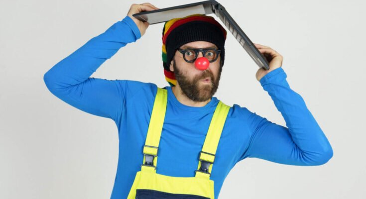A clown in bright clothes holds a laptop above his head.