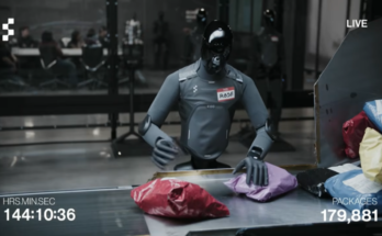 A humanoid robot with a gray body and black, reflective head stands next to a package sorting station while moving boxes and padded envelopes onto a conveyor belt.