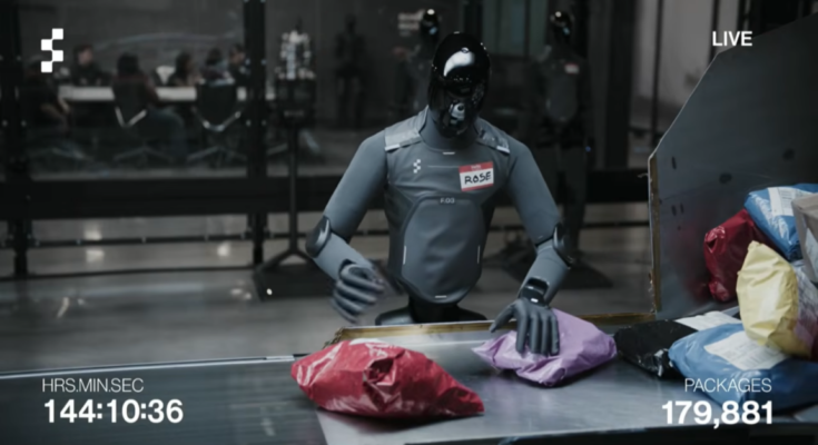 A humanoid robot with a gray body and black, reflective head stands next to a package sorting station while moving boxes and padded envelopes onto a conveyor belt.