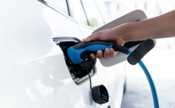 A parking lot with charging stations for electric cars. A hand holds a charging handle while charging an electric car at a home charging station.