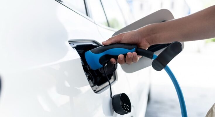 A parking lot with charging stations for electric cars. A hand holds a charging handle while charging an electric car at a home charging station.