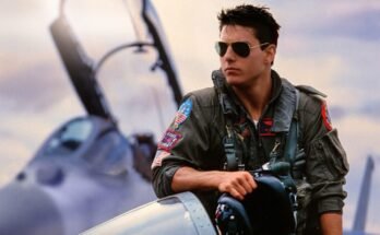 young to cruise in fighter pilot uniform and sunglasses casually leaning against a fighter jet