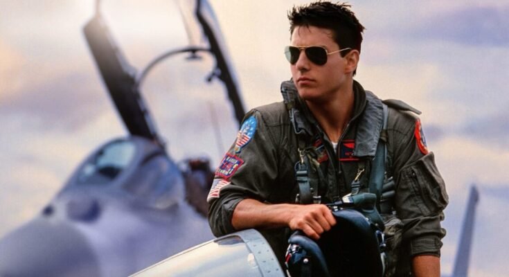 young to cruise in fighter pilot uniform and sunglasses casually leaning against a fighter jet
