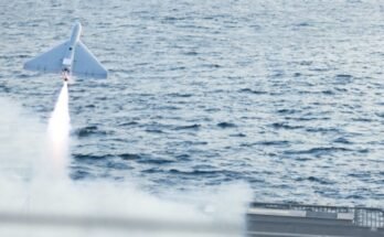 A military drone takes off from the flight deck of a combat ship. A rectangular satellite dish can be seen on the drone's top side.