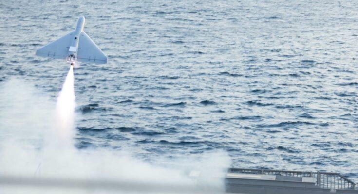A military drone takes off from the flight deck of a combat ship. A rectangular satellite dish can be seen on the drone's top side.