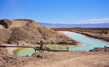 Image of a dry, plant less desert area. In amidst large lumps of soil sit a human-shaped pond of vivid blue water.