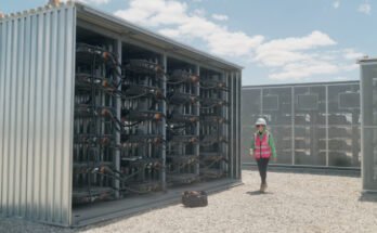A woman wearing a white safety helmet and a red safety vest is walking by a large steel structure containing used EV battery packs connected together. Such repurposed EV batteries provide 32 megawatt-hours of energy storage to support the local grid in Lancaster, California.