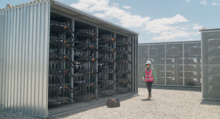 A woman wearing a white safety helmet and a red safety vest is walking by a large steel structure containing used EV battery packs connected together. Such repurposed EV batteries provide 32 megawatt-hours of energy storage to support the local grid in Lancaster, California.