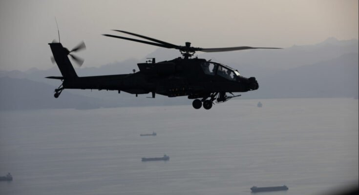 A US Army AH-64 Apache helicopter gunship can be seen flying over commercial ships in the Strait of Hormuz during a patrol on April 17, 2026.