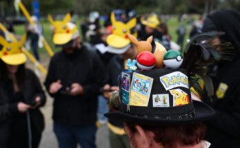 A player wearing a hat decorated with Pokemon characters and trading cards plays Pokemon GO on a smartphone during the in-person Pokemon GO Tour: Kalos Los Angeles 2026 event at the Rose Bowl Stadium in Pasadena, California, on February 20, 2026.