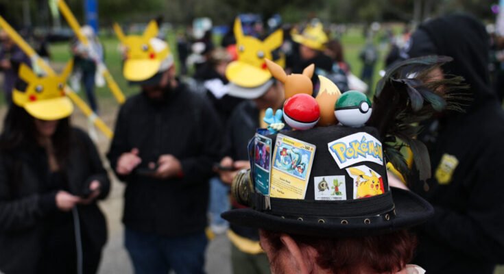 A player wearing a hat decorated with Pokemon characters and trading cards plays Pokemon GO on a smartphone during the in-person Pokemon GO Tour: Kalos Los Angeles 2026 event at the Rose Bowl Stadium in Pasadena, California, on February 20, 2026.