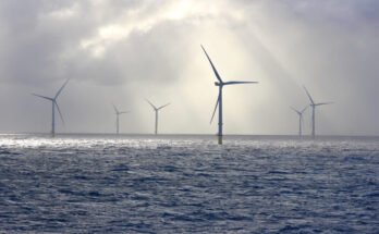 A series of tall wind turbines in the ocean, illuminated by beams of sunlight filtering through baps of heavy clouds.
