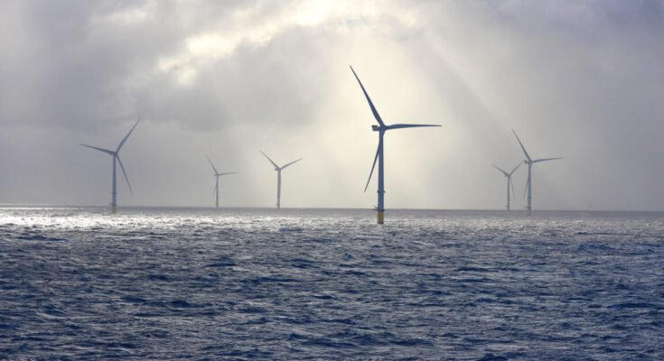 A series of tall wind turbines in the ocean, illuminated by beams of sunlight filtering through baps of heavy clouds.