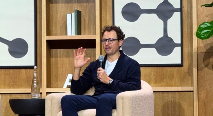 Dario Amodei sits on a chair on stage and gestures as he talks.