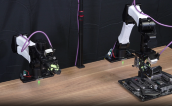 Two robotic arms sit on a lab table, with the robotic arm on the right holding a GPU above a motherboard. Teams of AI coding agents can train robots to do various manipulation tasks.
