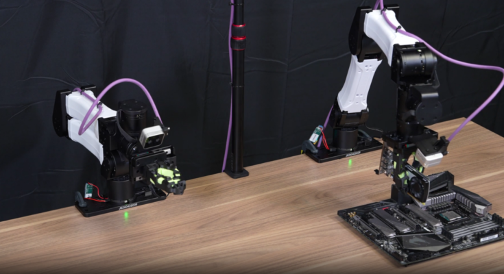 Two robotic arms sit on a lab table, with the robotic arm on the right holding a GPU above a motherboard. Teams of AI coding agents can train robots to do various manipulation tasks.