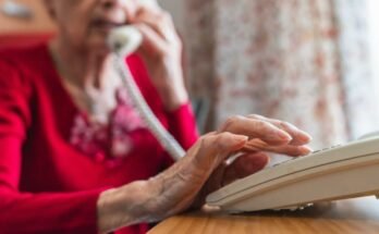 Elderly woman sitting at a table and dialing a phone.