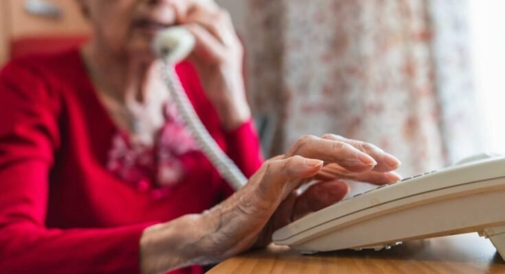 Elderly woman sitting at a table and dialing a phone.